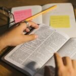 Close-up of hands holding and reading a Bible with study notes and a pencil on a desk.